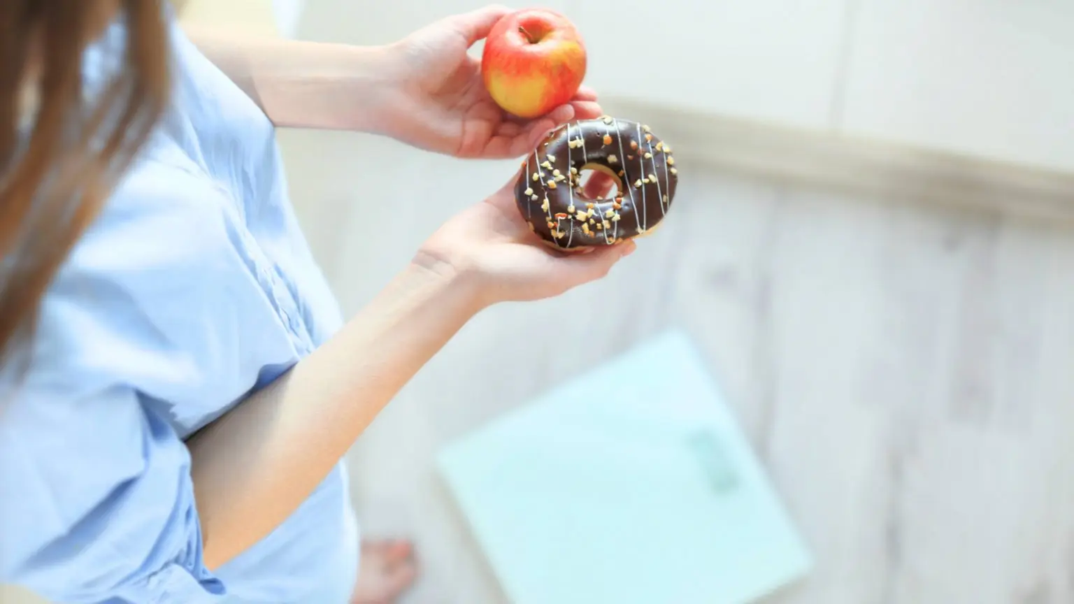 a-woman-holding-an-apple-in-one-hand-a-sprinkled-donut-in-the-other-while-standing-near-a-scale-to-weigh-herself-16x9-1-1536x864.webp